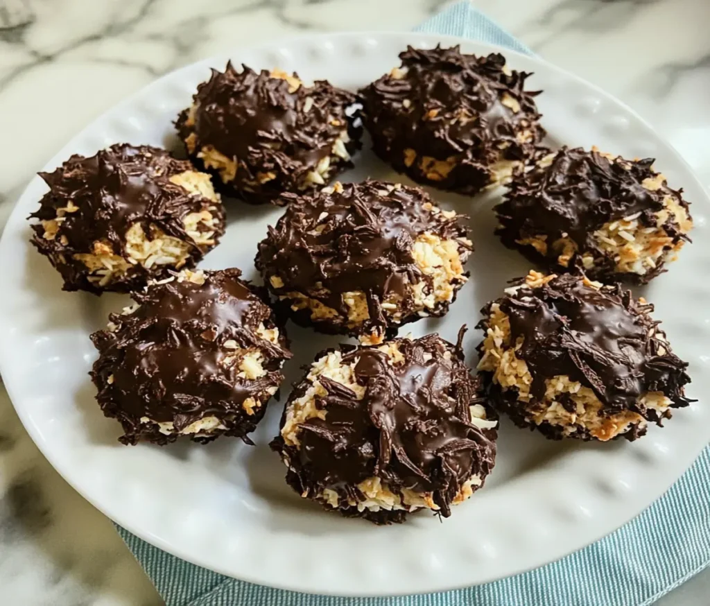 Close-up of freshly baked Schokoladen-Kokos-Makronen, golden-brown coconut macaroons dipped in glossy dark chocolate, arranged on a festive white plate with a holiday-themed background.